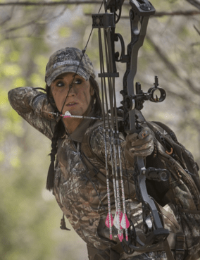 A woman in hunting gear takes careful aim with her compound bow